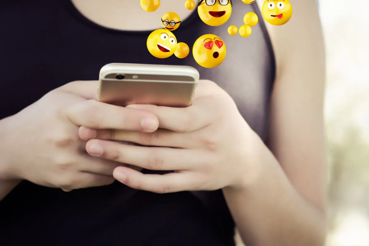 Pessoa segura um iPhone dourado com as duas mãos enquanto emojis de rosto, incluindo olhos de coração, saem da tela