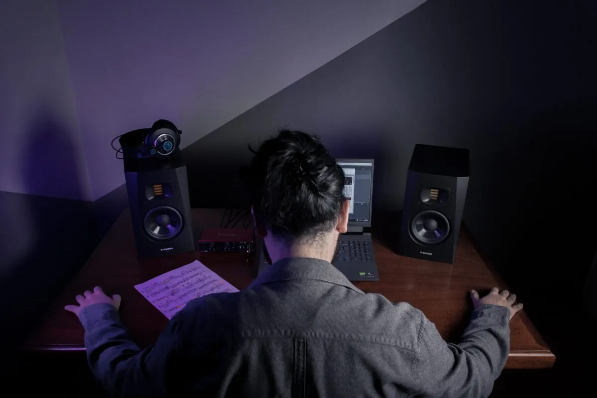 Pessoa produzindo música em um home studio, com laptop, monitores de estúdio e partitura sobre a mesa