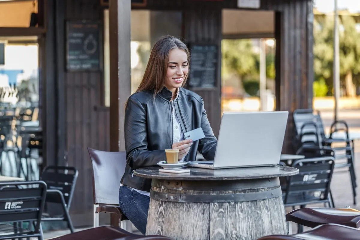 Mulher sentada em mesa de café, usando laptop e segurando cartão de crédito
